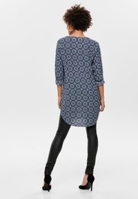 Tunic blu navy con motivo circolare, maniche a tre quarti e orlo arrotondato, abbinata a pantaloni in ecopelle neri e tacchi neri.