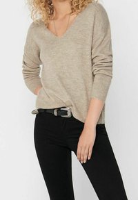 Beżowy sweter w serek z miękkiej, textured materiału z długimi rękawami, w połączeniu z dopasowanymi czarnymi dżinsami i czarnym paskiem z srebrną klamrą.