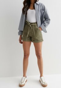 Pantaloni corti verde oliva con vita regolabile; abbinati a una canotta bianca e a una camicia button-up a righe blu. Sneakers bianche con dettagli beige.