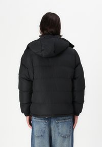 Μαύρο πούφφερ μπουφάν με αποσπώμενη κουκούλα, διαθέτοντας επενδυμένο σχέδιο και ελαστικές μανσέτες. Φορεμένο πάνω από ανοιχτού μπλε τζιν παντελόνι.