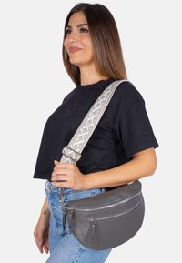 Sac en cuir gris en bandoulière avec une forme courbée, deux poches zippées et une sangle ajustable à motif. Surface texturée et quincaillerie en argent.