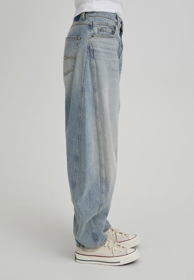 Gaiši zilas oversize džinsa bikses ar vaļīgu piegriezumu, nevienmērīgu izbalēšanu un divām aizmugurējām kabatām, kombinētas ar krēmkrāsas auduma sporta apaviem.