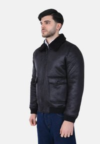 Uomo che indossa una giacca bomber nera con collo in shearling, dotata di tasche frontali e jeans blu, in piedi contro uno sfondo chiaro e semplice.