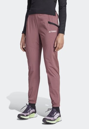 Personne portant un pantalon d'extérieur mauve avec des poignets élastiques et une poche zippée sur la cuisse, associé à des chaussures de sport noires et violettes.