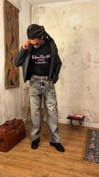 Mand med dreadlocks, der justerer solbriller, iført læderjakke, grafik t-shirt, syre-vaskede jeans, står i et rustikt rum med tæppe og kuffert.