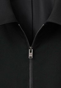 Črni žep z zadrgo z gladko teksturo tkanine; vsebuje srebrno zadrgo z obeskom z blagovno znamko in stoječi ovratnik.