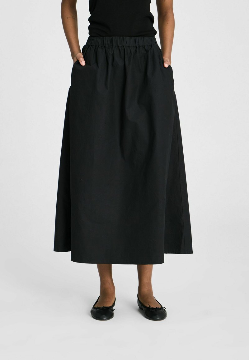 Personne portant un haut noir sans manches, une longue jupe noire élastique à la taille avec poches, et des ballerines noires, debout sur un fond uni.