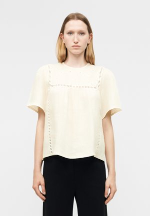 Γυναίκα με ίσια ξανθά μαλλιά φοράει μια άνετη κρεμ μπλούζα με λεπτομέρειες eyelet και μαύρο παντελόνι, όρθια μπροστά σε λευκό φόντο.