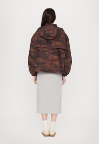 Braune Tarnjacke in Übergröße mit Kapuze, Fronttasche und elastischen Bündchen, getragen über einem grauen knielangen Rock und beigen Schuhen.
