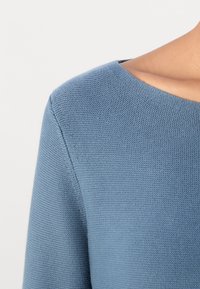 Gros plan sur une personne portant un pull en maille texturée bleu clair avec un col rond sur fond uni.