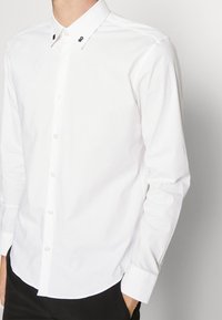 Chemise blanche à manches longues en coton avec col classique, dotée de boutons noirs en accent et d'un ourlet arrondi.