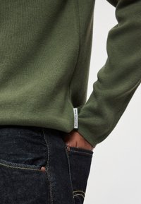 Camisola verde-oliva com tecido texturizado, design ajustado e etiqueta lateral. Combinada com calças de ganga escuras com detalhes em cobre.