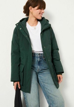 Dunkelgrüne isolierte Jacke mit Kapuze, vorderer Druckknopfverschluss und großen Seitentaschen, kombiniert mit lockeren Jeans. Lässiger Stil.