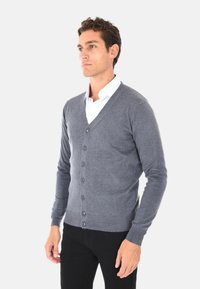 Cardigan grigio realizzato in tessuto morbido, con scollo a V, sei bottoni sul davanti e polsini a coste. Indossato sopra una camicia bianca con colletto.