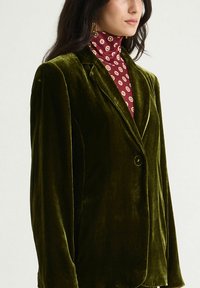 Blazer vert en velours avec revers en encoche et une fermeture à bouton unique, doté d'épaules structurées et d'une texture lisse.