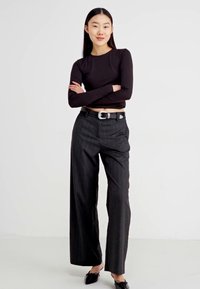 Top negru cu maneci lungi, pantaloni cărbune cu dungi verticale, talie înaltă, croială largă, cataramă argintie la curea, loaferi negri. Fundal alb simplu.