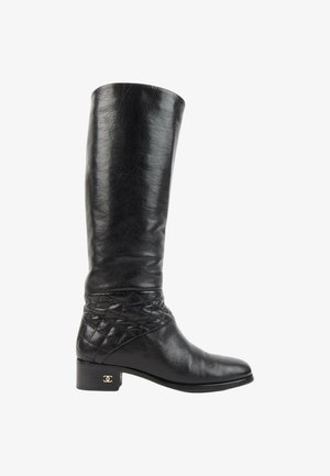 Botte noire en cuir montante jusqu'au genou avec un petit talon, détail matelassé autour de la cheville et un petit logo doré sur le talon.