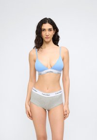 Γυναίκα με σγουρά μαλλιά, φορώντας ανοιχτό μπλε σουτιέν και γκρι εσώρουχα Calvin Klein, στέκεται μπροστά σε έναν απλό λευκό φόντο.