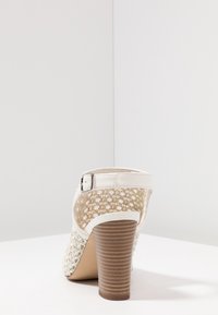 Lodičkové boty s tkaným béžovým zadním dílem, bílým lakovaným páskem a hnědým vroubkovaným podpatkem. Obsahují sponu pro bezpečné zapínání.