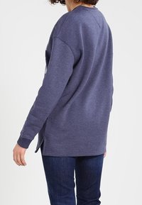 Felpa viola con vestibilità ampia, scollo rotondo e maniche lunghe. Presenta spaccature laterali e polsini a costine, abbinata a jeans blu.