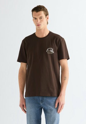 Młody mężczyzna z krótkimi włosami, ubrany w ciemnobrązowy t-shirt Wrangler Denim Club i niebieskie jeansy, stojący na jasnym, jednolitym tle.