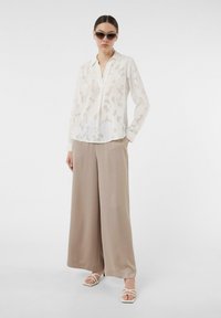 Doorzichtig wit blouse met een subtiel patroon, gecombineerd met beige wijde broek en witte sandaaltjes met bandjes. Model draagt een donkere zonnebril.