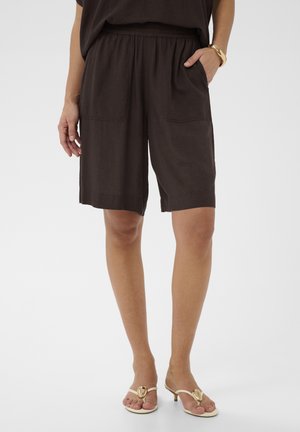 Femme portant un short ample marron foncé arrivant aux genoux avec des poches avant, associé à des sandales blanches à talons avec des accents dorés.