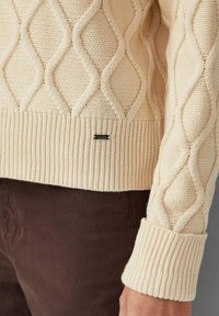 Beiger, kabelgestrickter Pullover mit geripptem Saum und Bündchen. Strukturiertes Rautenmuster an den Ärmeln. Kleines schwarzes Markenetikett nahe dem Saum.