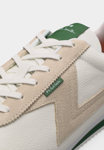 Baskets blanches et beiges avec lacets blancs, accents verts sur la semelle, et étiquettes de marque vertes portant l'inscription "PS Paul Smith" sur le côté et la languette.