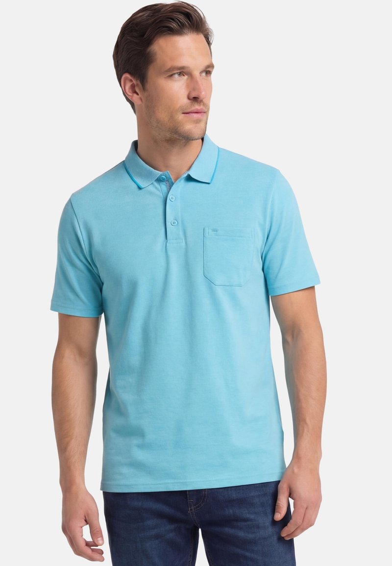 Homme portant un polo à manches courtes bleu clair avec une poche poitrine et un jean bleu foncé, debout et regardant vers sa droite.