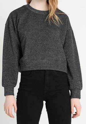 Woman wearing a dark gray long-sleeve cropped sweatshirt and high-waisted black pants against a plain background.