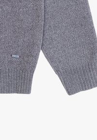 Grijze gebreide trui met een textuurpatroon, geribde manchetten en een klein blauw label met rode accenten aan de onderkant.