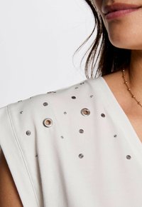 Witte top met zilveren accenten en decoratieve grommets op de schouder. Zacht stof met een losse pasvorm en een ronde halslijn.