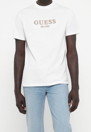 Homme portant un T-shirt blanc à manches courtes avec "GUESS EST. 1981" en lettres marron, associé à un jean bleu clair sur fond uni.