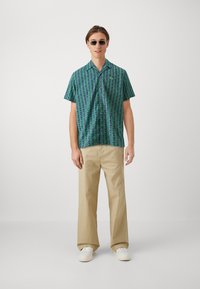 Cămașă cu mâneci scurte, cu un model geometric în verde și albastru, asortată cu pantaloni bej și adidași albi. Ochelari de soare purtați.