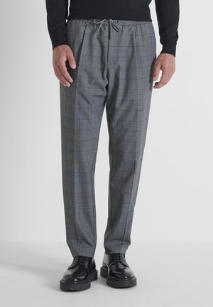Man in grijs geruite broek met een tailleband met koord en zwarte gepolijste veterschoenen die op een effen oppervlak staat.