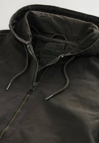 Jachetă cu capuciu de culoare maro închis, având un fermoar frontal, capuc cu șnur și o etichetă din material cu marca "STANDARD ISSUE".