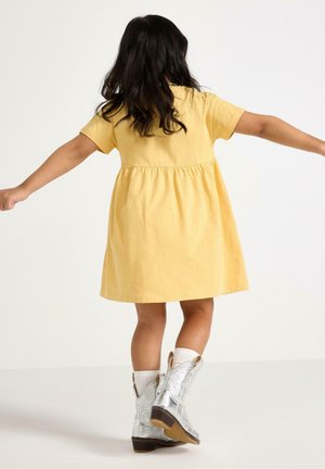 Barn med langt mørkt hår iført gul kjole og sølvstøvler står med armene udstrakt mod en ensfarvet hvid baggrund.