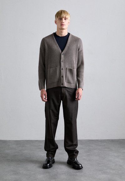 Cardigan côtelé marron clair avec deux poches avant, col en V et boutons, associé à un pantalon sombre et des chaussures noires, se tenant sur un sol gris.