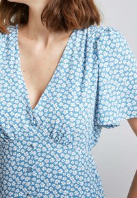Robe florale bleue avec des manches courtes bouffantes, un décolleté en V profond et des boutons décoratifs le long du corsage, fabriquée dans un tissu léger.