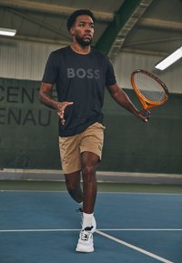 Fekete pólót viselő sportoló, rajta a "BOSS" felirattal, bézs rövidnadrágban és fekete akcentús fehér cipőben, narancssárga teniszütőt tartva egy pályán.