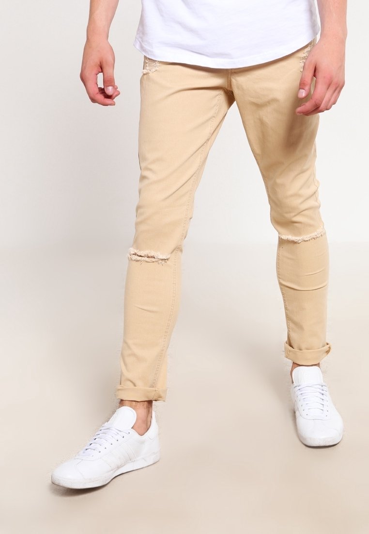 Pantaloni beige slim fit con ginocchia strappate, abbinati a sneaker bianche e una camicia bianca, indossati da una persona in piedi su uno sfondo chiaro.