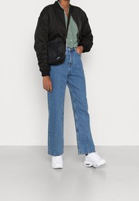Persona che indossa una giacca bomber nera, una camicia verde a bottoni, jeans blu, sneakers bianche e una borsa a tracolla nera Nike, in piedi con una mano in tasca.