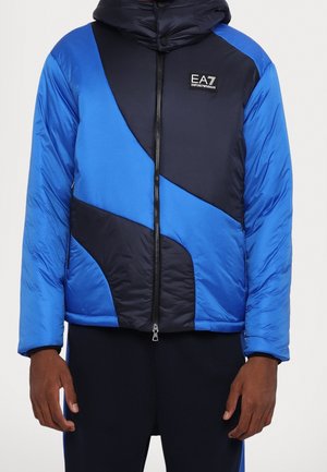 Mand iført en blå og sort polstret jakke med diagonale farveblokke og EA7 Emporio Armani-logo på brystet, stående med armene langs siden.