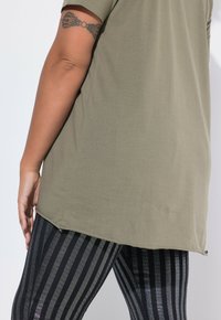 Top de manga corta color verde oliva con dobladillo curvado y leggings a rayas negras y plateadas con una textura brillante. Tatuaje visible en el brazo.