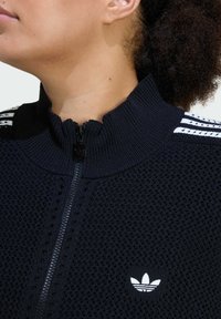 Čierna bunda na zips z textúrovanej pleteniny, s vysokým golierom a bielym logom adidas s tromi horizontálnymi pruhmi na pleciach.