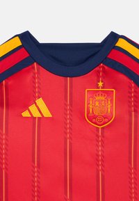 Camisa desportiva vermelha feita de poliéster, com riscas amarelas nos ombros, um logótipo da Adidas em amarelo bordado e o emblema de Espanha com uma estrela dourada.