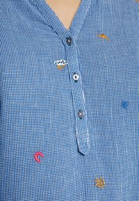 Chemise à carreaux bleue avec de petites icônes brodées comprenant un éclair, un nuage avec de la pluie, une étoile, un soleil et des formes de fer à cheval rouges.