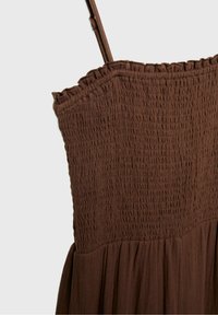 Robe marron sans manches avec corsage smocké, fines bretelles et détail à volants au niveau de l'encolure sur un fond uni.