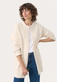 Knitted cream cardigan featuring a textured pattern, open front, and two front pockets, paired with a white t-shirt and blue jeans.